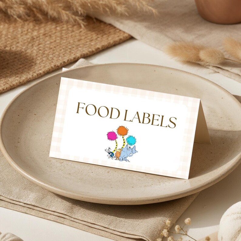 May include: A folded food label card with the words "FOOD LABELS" in brown. The card features a light beige gingham pattern border and a colorful illustration of characters and trees. The card is placed on a beige plate.
