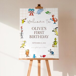May include: A wooden easel displays a welcome sign for Olive's first birthday. The sign features a cream-colored background with a light checkered pattern and colorful cartoon characters. Text reads "Welcome To Olive's First Birthday, September 21, 2026".