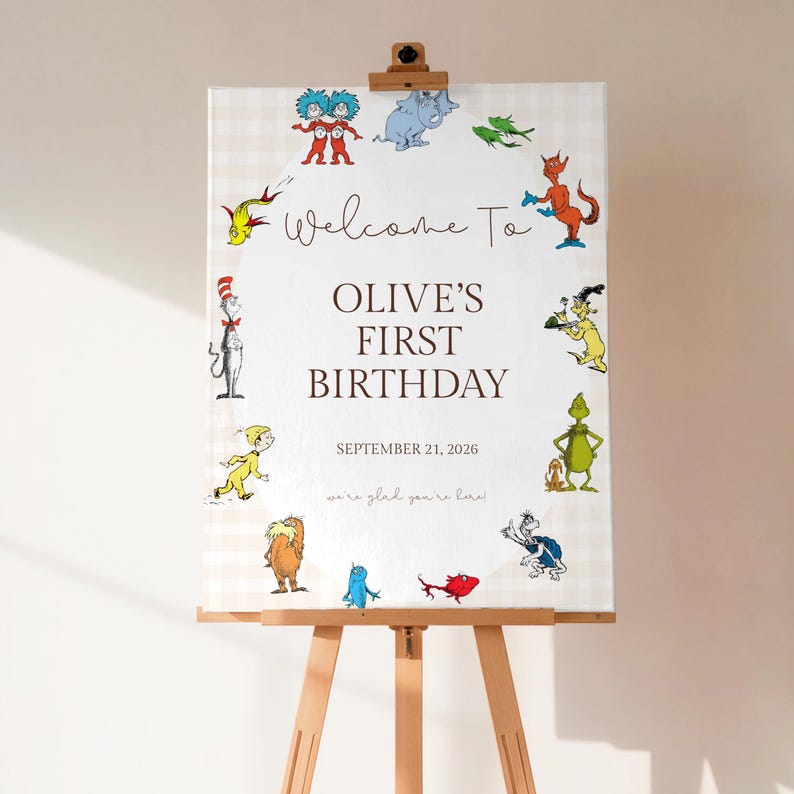 May include: A wooden easel displays a welcome sign for Olive's first birthday. The sign features a cream-colored background with a light checkered pattern and colorful cartoon characters. Text reads "Welcome To Olive's First Birthday, September 21, 2026".