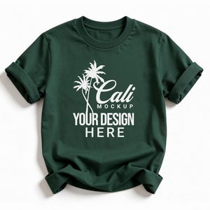 May include: Dark green t-shirt with rolled sleeves. The shirt features a white graphic of palm trees and the text "Cali Mockup YOUR DESIGN HERE". The t-shirt is a mockup, perfect for showcasing designs.