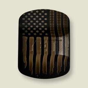 Puede incluir: Objeto rectangular negro con un diseño de bandera estadounidense desgastada. La bandera presenta estrellas doradas y rayas marrones, con una fina línea azul. El objeto tiene bordes superior e inferior ligeramente redondeados.