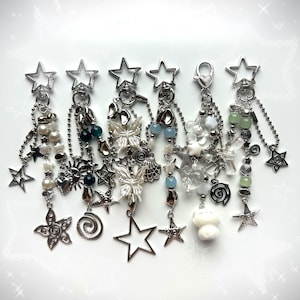 May include: Six silver-toned metal keychains featuring star-shaped clasps and various dangling charms including butterflies, hearts, flowers, seashells, suns, and beads in shades of white, blue, and green, all suspended from silver ball chains.
