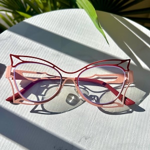 Puede incluir: Gafas rosas de ojo de gato con detalles rojos. Las monturas tienen un diseño único con bordes puntiagudos y un doble puente. Las lentes son transparentes y las gafas están sobre una superficie blanca con sombras y vegetación en el fondo.