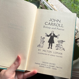 May include: An open book with the title "JOHN CARROLL Bishop and Patriot" in black text. The book's illustration depicts a figure with arms outstretched. The book is published by Vision Books, with the publisher's name and location printed below.
