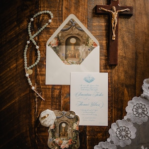 May include: A wedding invitation set featuring a white envelope with a floral and religious interior print, a card with blue calligraphy, a light green beaded rosary, a gold ring in a white box, a wooden crucifix, and a white lace veil.