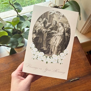 May include: A beige wedding card featuring a sepia-toned illustration of a marriage ceremony within an oval frame. White roses adorn the bottom, and the text "Blessings on Your Marriage" is written in elegant script.