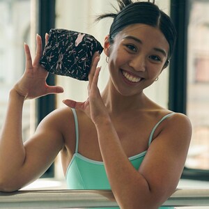 May include: A woman in a turquoise leotard and a pink and brown knitted scarf is smiling and holding a black and pink patterned pouch in front of her face. She is standing in a dance studio with a barre behind her.