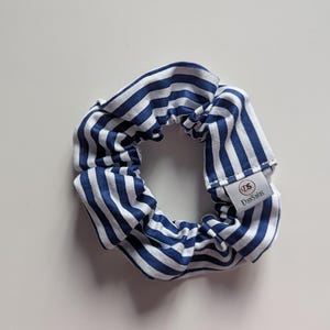 May include: A blue and white striped scrunchie. The fabric is gathered and elasticized, creating a ruffled effect. A small white tag with black text is attached to the scrunchie. The scrunchie is a hair accessory.