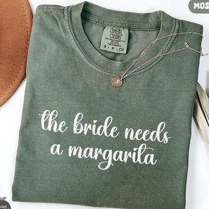 May include: A moss green t-shirt with the phrase "the bride needs a margarita" in white script. A gold necklace with a pendant rests on the shirt. The shirt is folded and displayed with a brown hat and decorative elements.