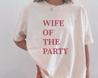 May include: Ivory-colored t-shirt with the text "WIFE OF THE PARTY" in a bold, red-pink font. The shirt has short sleeves and a crew neck. The bottom of the shirt is partially visible, along with a red and white checkered pattern.