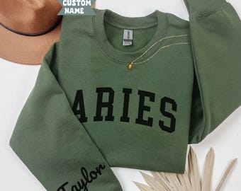 Op de afbeelding: Militairgroen Aries sweatshirt met zwarte blokletters. Het woord "Taylor" is in cursief op de linkermouw gedrukt. Een gouden ketting en een bruine hoed zijn zichtbaar op de achtergrond. De sweatshirt is gemaakt van een zacht materiaal.