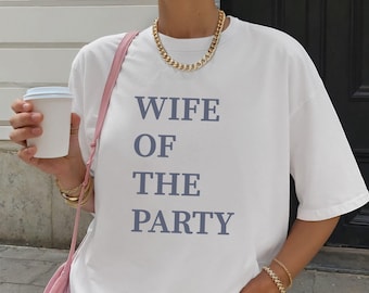 May include: White t-shirt with the text "WIFE OF THE PARTY" in blue. The shirt is a casual style, with a relaxed fit. The model is wearing gold jewelry and holding a white coffee cup.