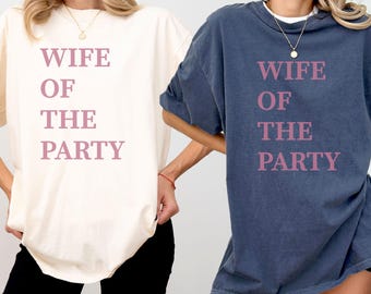 May include: Two t-shirts, one ivory and one denim blue, each with the text "WIFE OF THE PARTY" in a pink font. The ivory shirt is paired with black shorts. The denim shirt is paired with black shorts.