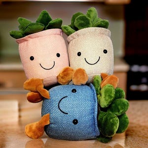 May include: Three plush toy succulents in pots. The succulents are green and the pots are pink, beige, and blue. Each pot has a smiling face.