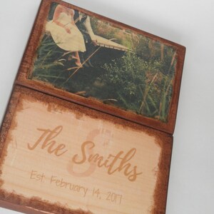May include: Wooden hinged photo box with a rustic brown frame. The top section displays a photo of a couple near a body of water. The bottom section has the text "The Smiths" and the date "Est. February 14, 2017".