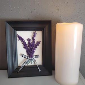 May include: A framed artwork featuring a purple lavender bouquet tied with a black and white checkered ribbon. The artwork is in a black frame. A large white candle is next to the framed art.