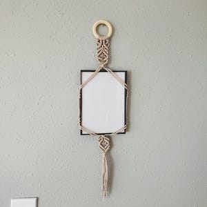 May include: A macrame wall hanging with a black framed picture holder. The macrame is beige and features a wooden ring at the top and a decorative knot with a tassel at the bottom. The wall is painted a light gray color.