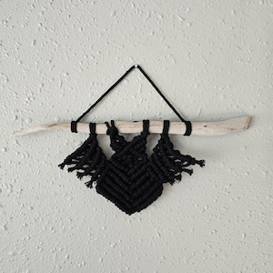 Könnte beinhalten: Schwarzer Makramee-Wandbehang in Fledermaus-Design. Der Makramee-Wandbehang ist an einem Stück Treibholz und einer schwarzen Schnur befestigt. Die Fledermaus hat einen rautenförmigen Körper und gefranste Flügel. Der Hintergrund ist eine strukturierte weiße Wand.