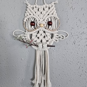 May include: A white macrame owl wall hanging. The owl features wooden bead eyes, gold hoop accents, and a small branch with pink flowers. The owl is suspended by a white cord and has a fringe at the bottom.