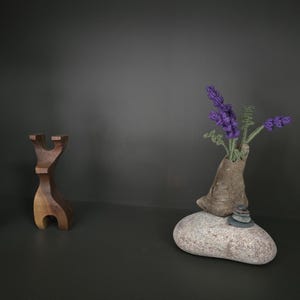 May include: Two abstract wooden sculptures with smooth, curved forms stand beside a textured stone base. A small, tan, organic-shaped vessel sits on the stone, holding several sprigs of purple lavender flowers with green stems and leaves.