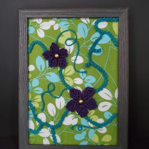 May include: A framed artwork featuring a green floral fabric background. Two purple crocheted flowers with gold centers are placed on the fabric. Teal yarn vines and a gray wooden frame complete the piece.