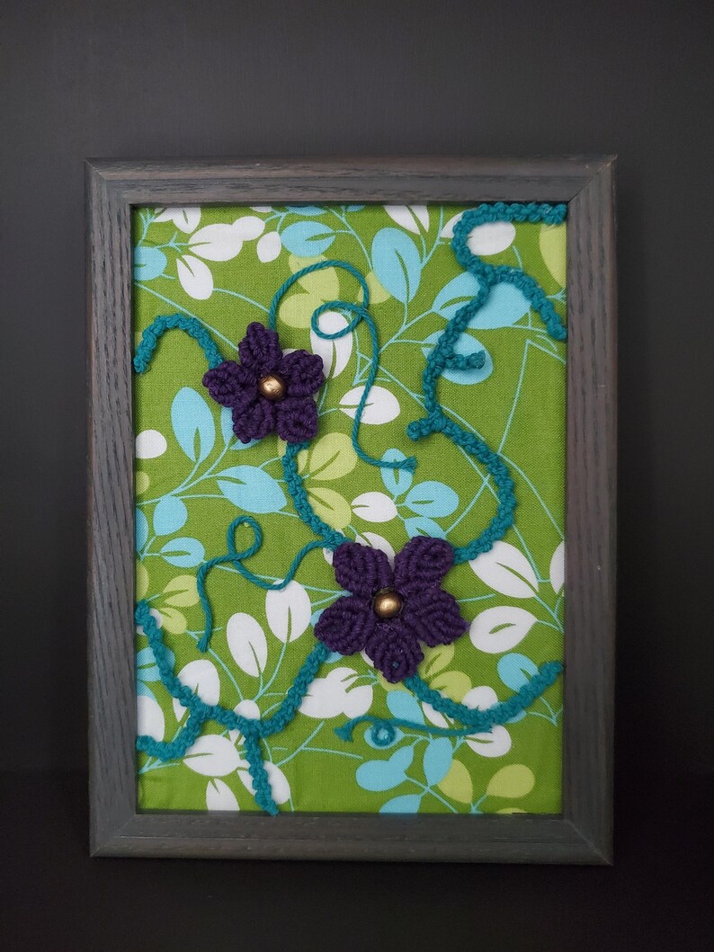 May include: A framed artwork featuring a green floral fabric background. Two purple crocheted flowers with gold centers are placed on the fabric. Teal yarn vines and a gray wooden frame complete the piece.