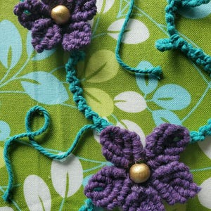 May include: Close-up of two handmade crochet flowers in purple with gold centers, connected by turquoise yarn. The flowers are set against a green and white floral patterned fabric, showcasing a handcrafted floral design.