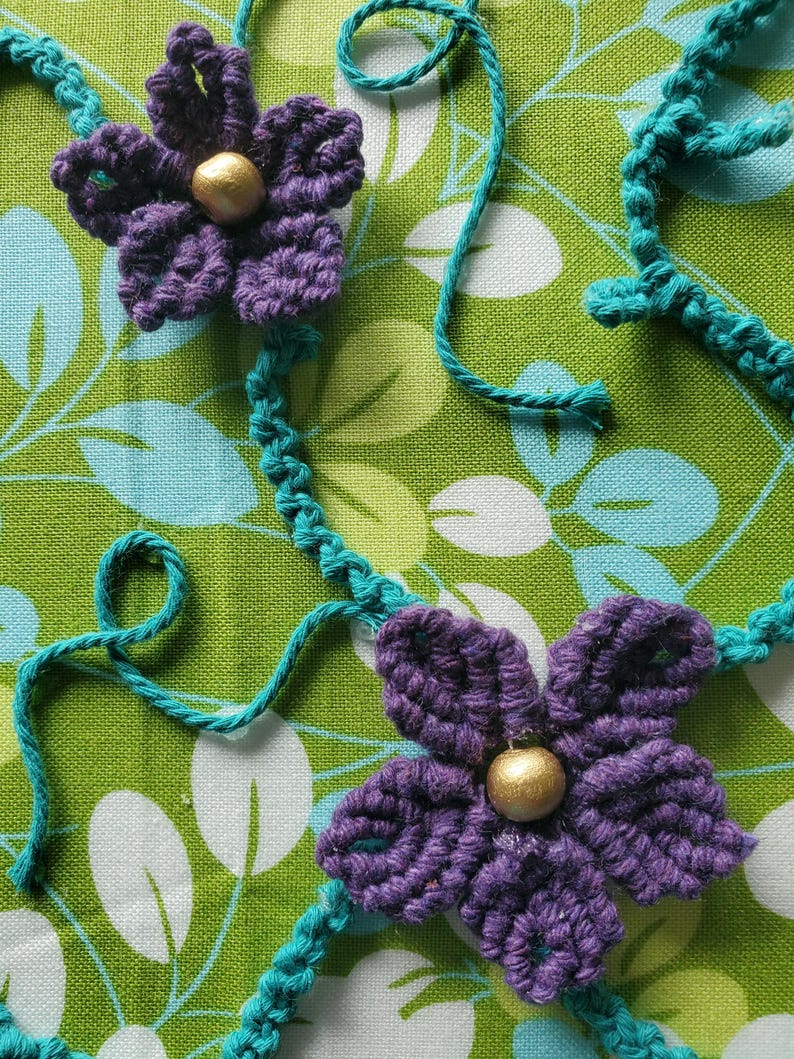 May include: Close-up of two handmade crochet flowers in purple with gold centers, connected by turquoise yarn. The flowers are set against a green and white floral patterned fabric, showcasing a handcrafted floral design.