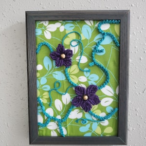 May include: Framed artwork featuring a green floral fabric background with white and blue leaves. Two purple crocheted flowers with gold centers are accented by turquoise crocheted vines. The frame is dark gray.