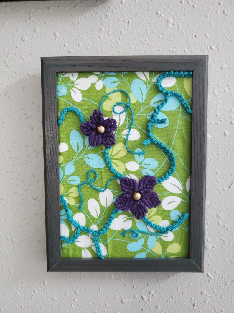 May include: Framed artwork featuring a green floral fabric background with white and blue leaves. Two purple crocheted flowers with gold centers are accented by turquoise crocheted vines. The frame is dark gray.
