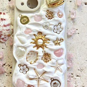 Op de afbeelding: Een witte telefoonhoes met een getextureerd, romig oppervlak, versierd met gouden bedels, roze edelstenen en zeesterren. De bedels omvatten een hartvormig medaillon, bloemen en een zonnestraal. De tekst "Quartz Cove" staat onderaan.