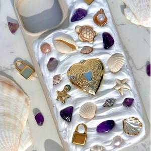 May include: A beige phone case decorated with white resin, seashells, amethyst stones, and gold charms, including a heart-shaped locket, starfish, and padlocks. The case is surrounded by additional seashells and amethyst crystals.