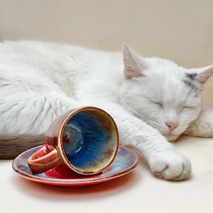 Puede incluir: Un juego de taza y platillo de cerámica con un esmalte brillante, que presenta un interior azul profundo que se transforma en tonos marrón rojizo en el exterior y el borde. La taza tiene un asa curva y descansa sobre un platillo redondo a juego.