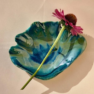Könnte beinhalten: Eine glänzende, unregelmäßig geformte Keramikschale in Muscheloptik mit einem Farbverlauf aus tiefem Petrol, Türkis und Limettengrün. Eine einzelne rosa Sonnenhutblüte mit dunkelbrauner Mitte und langem grünen Stiel liegt diagonal auf der Schale.