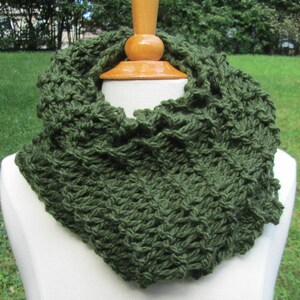 May include: A dark green, hand-knitted infinity scarf. The scarf is displayed on a white mannequin head and neck, showcasing the textured stitch pattern. The background is a blurred view of green grass and trees.