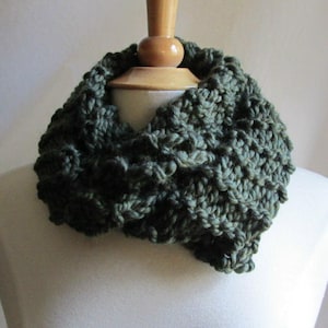 May include: A dark green, chunky knit cowl scarf. The scarf is made of a thick, textured yarn, creating a warm and cozy appearance. The cowl is displayed on a mannequin, showcasing its circular design and potential for winter wear.