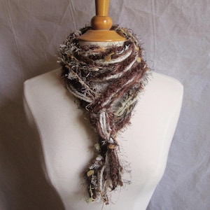 May include: A handmade, fringed scarf in shades of brown, cream, and olive green, displayed on a mannequin. The scarf features a textured, layered design with a knotted end, showcasing a bohemian style.