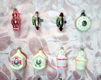 May include: A collection of vintage Christmas ornaments. The ornaments include a pink robot, rocket ships, a clock, cat-shaped ornaments, and house-shaped ornaments with a cat inside. The ornaments are made of glass and have silver hanging loops.