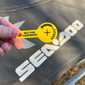 Puede incluir: Una herramienta de pl&aacute;stico amarilla con el texto "NOT FOR TIGHTENING" y "LOOSEN" con una flecha. La herramienta se sostiene en una mano, con el logotipo de Sea Doo en el fondo.