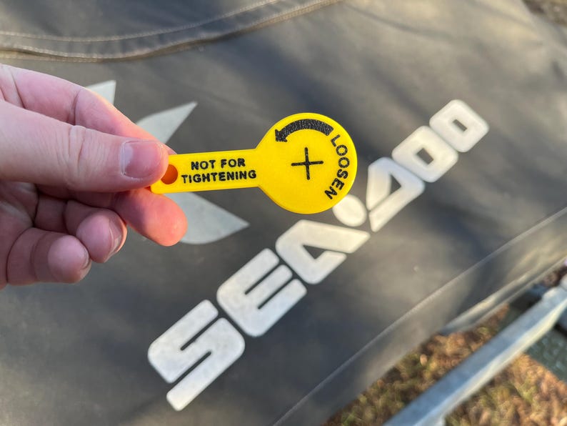Puede incluir: Una herramienta de pl&aacute;stico amarilla con el texto "NOT FOR TIGHTENING" y "LOOSEN" con una flecha. La herramienta se sostiene en una mano, con el logotipo de Sea Doo en el fondo.