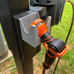 May include: A gray plastic hose holder mounted on a black wooden post. An orange and black garden hose nozzle is secured in the holder. The nozzle has the text "WATER MIST" printed on it. A black hose extends from the nozzle.