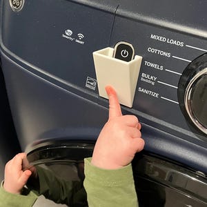 May include: A close-up of a dark blue washing machine control panel with white text labels for wash cycles. A small white container holds a black power button. A child's hand is touching the power button.