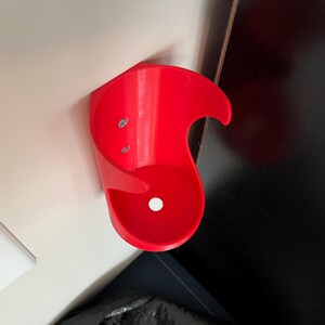 May include: A bright red plastic wall mount cup holder with two screw holes. The holder is designed to hold a single cup or bottle.