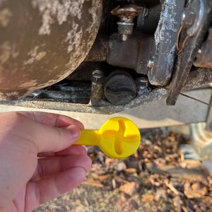 Puede incluir: Una herramienta amarilla con una cabeza en forma de cruz se sostiene cerca de una tapa negra en un componente met&aacute;lico. La herramienta parece ser utilizada para el mantenimiento o reparaci&oacute;n de maquinaria, posiblemente un barco o remolque.