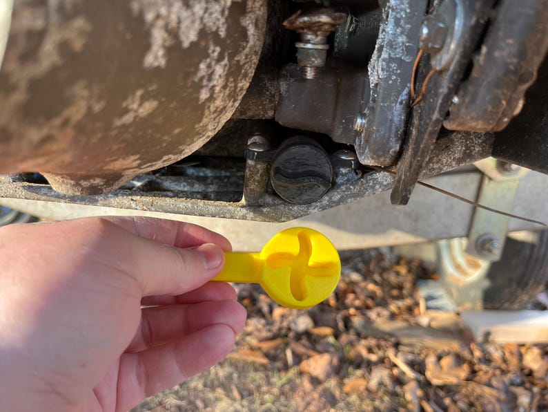 Puede incluir: Una herramienta amarilla con una cabeza en forma de cruz se sostiene cerca de una tapa negra en un componente met&aacute;lico. La herramienta parece ser utilizada para el mantenimiento o reparaci&oacute;n de maquinaria, posiblemente un barco o remolque.