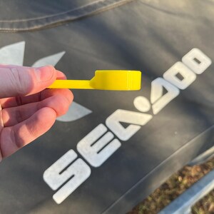 Puede incluir: Una pieza de pl&aacute;stico amarillo brillante con una cabeza rectangular y un mango fino y plano. La pieza se sostiene en una mano, con el fondo que muestra una superficie gris oscuro con el texto blanco "SEA 400".