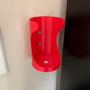 May include: A red plastic wall-mounted holder for a beverage can or bottle. The holder is attached to the wall with two screws.
