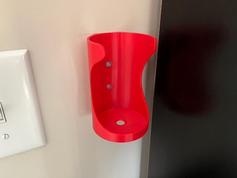 May include: A red plastic wall-mounted holder for a beverage can or bottle. The holder is attached to the wall with two screws.