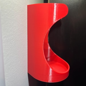 May include: A bright red plastic wall mount with a curved opening, designed to hold a small object like a toothbrush or a razor.