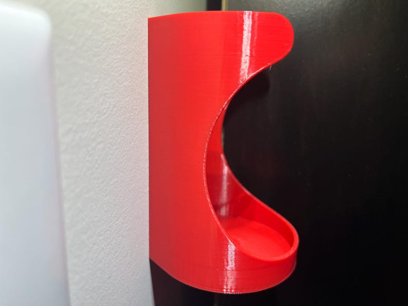 May include: A bright red plastic wall mount with a curved opening, designed to hold a small object like a toothbrush or a razor.